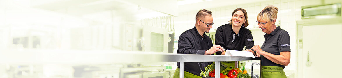 Karriere bei Dussmann: Gastronomie. Gruppenbild von drei lächelnden Dussmann-Mitarbeitenden aus dem Gastronomie-Bereich, die in einer Industrieküche stehen und gemeinsam ein Dokument anschauen.