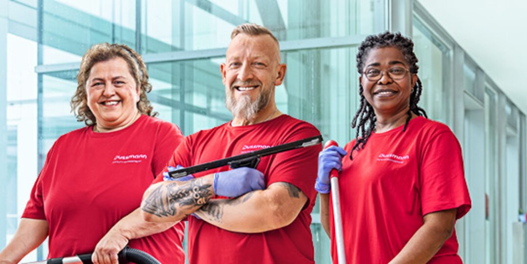 Gruppenbild von drei lächelnden Dussmann-Mitarbeitenden aus dem Arbeitsbereich Gebäudereinigung vor blauem Hintergrund. 