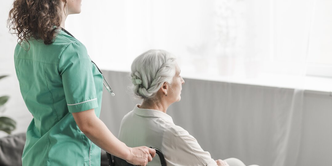 Dussmann Wäscherei-Service für Pflegeheime, der saubere, weiche und hygienische Wäsche liefert, die auf die Bedürfnisse der Bewohner zugeschnitten ist, und dabei strenge Hygieneprotokolle für das Wohlbefinden älterer Menschen einhält. | © <a href="https://www.freepik.com/free-photo/side-view-nurse-pushing-disabled-patient-wheel-chair_2652850.htm">Image by freepik</a>